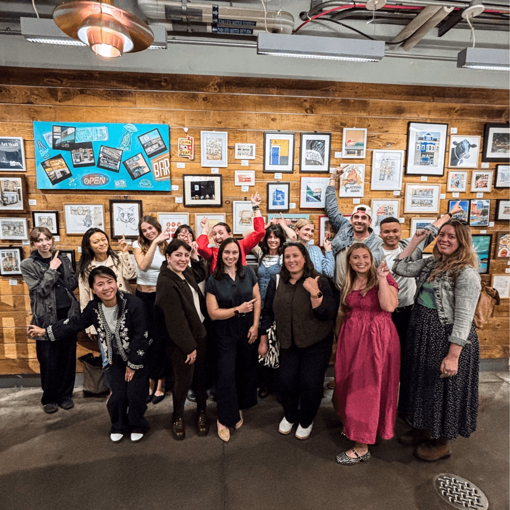 A group of smiling artists standing in front of a large gallery wall of original artwork in the Boston Public Market